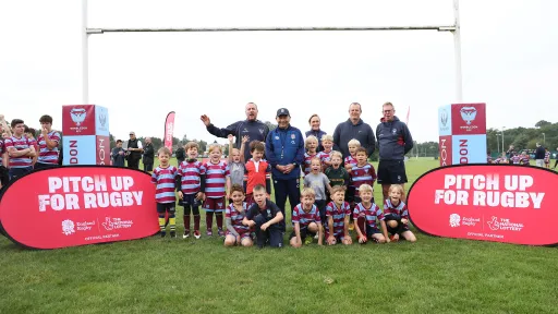 Group of children and adults posing on a rugby field with banners promoting "Pitch Up For Rugby."